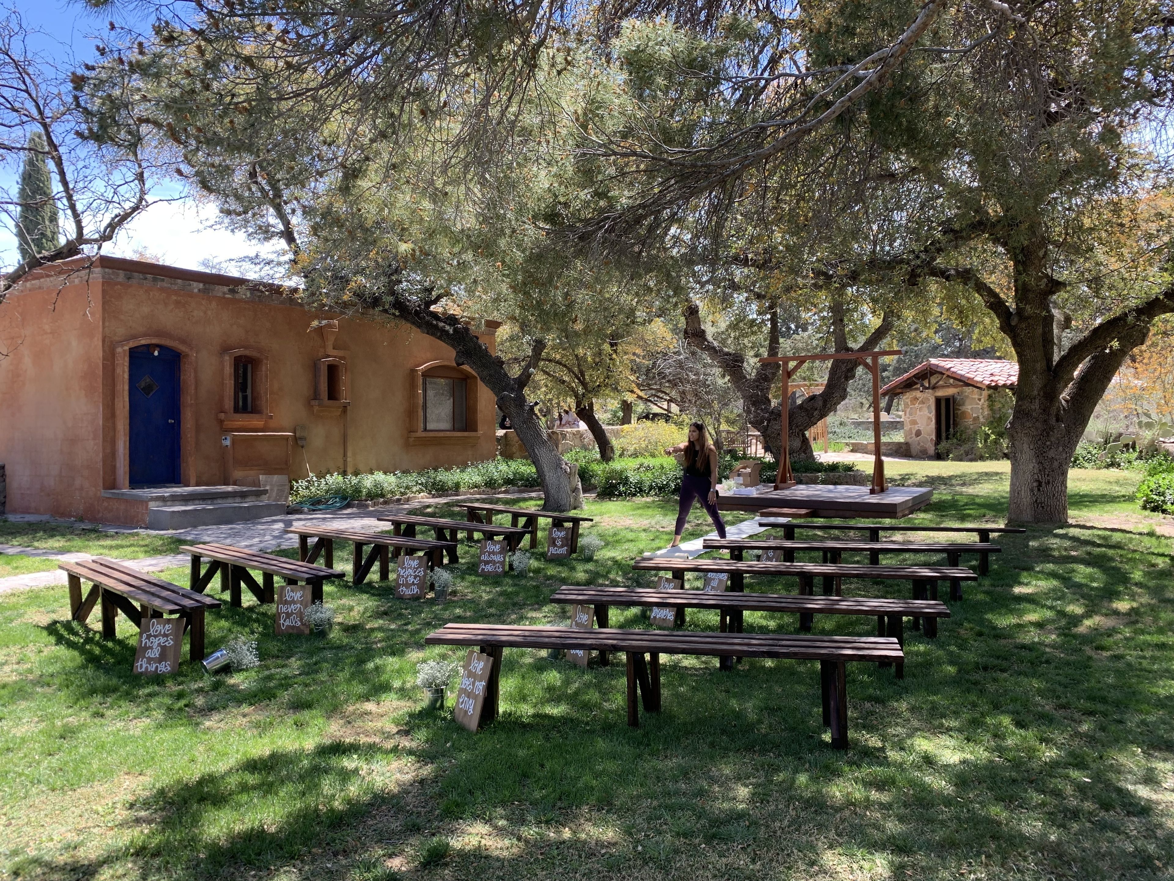 An outdoor area with picnic tables and benches, surrounded by trees and a rustic building with a blue door.