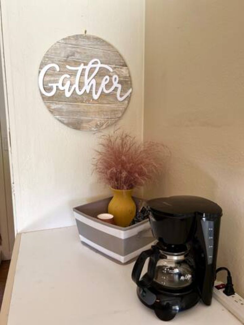 Een koffiezetapparaat op een aanrecht, met een decoratieve vaas en een bord met een tekst op de muur.