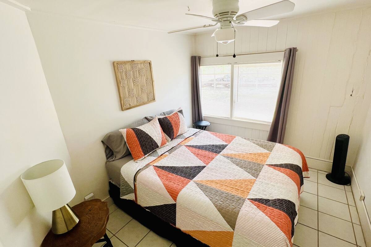 Una cama bien tendida con una colcha de estampado geométrico, una mesita de noche con lámpara, un ventilador de techo y una ventana con cortinas.