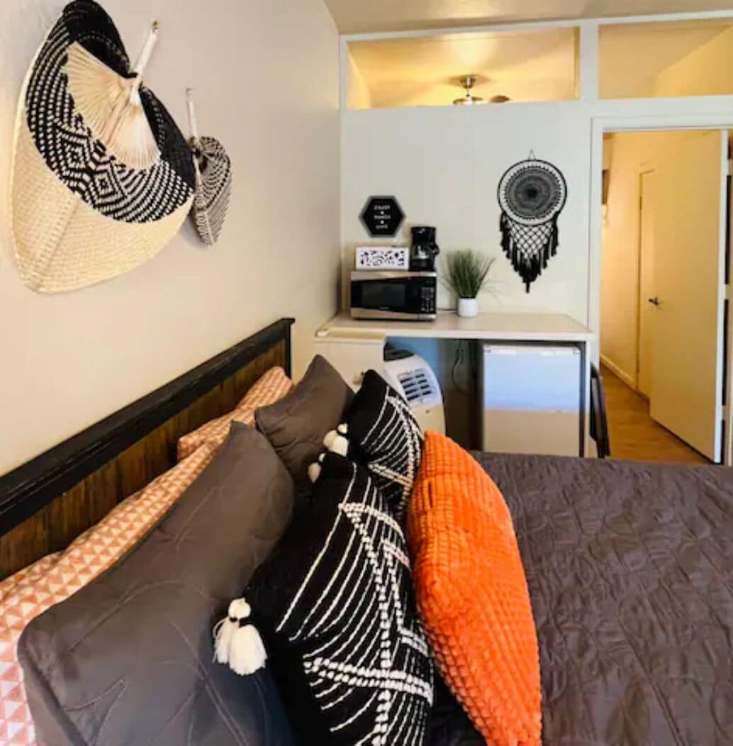 Un dormitorio con cama, almohadas, una mesita de noche con microondas, una planta y una pared decorada con un atrapasueños.