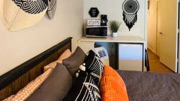 Un dormitorio con cama, almohadas, una mesita de noche con microondas, una planta y una pared decorada con un atrapasueños.