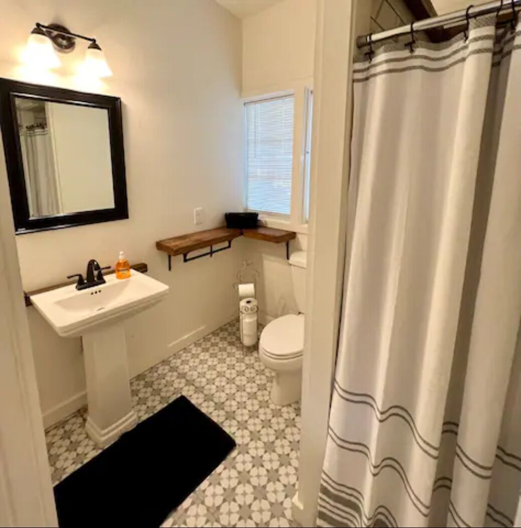 A small bathroom with a white sink, a mirror, a toilet, and a window with blinds.