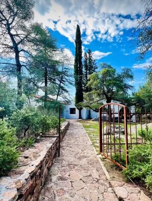 A stone pathway leading to a white building with a metal gate, surrounded by trees and greenery.