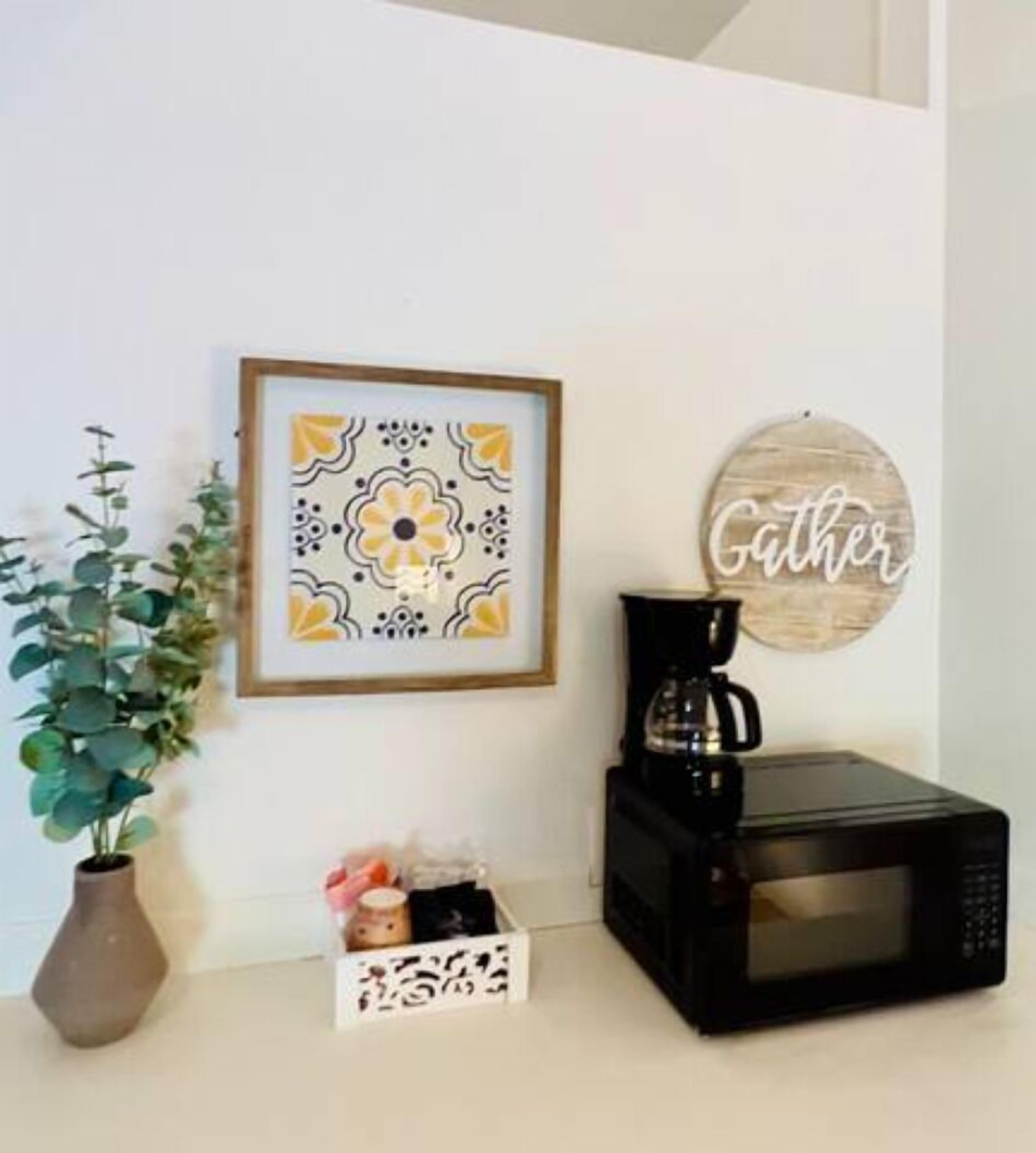 Una cocina con un cuadro de flores enmarco, una cafetera, un microondas y un letrero decorativo.
