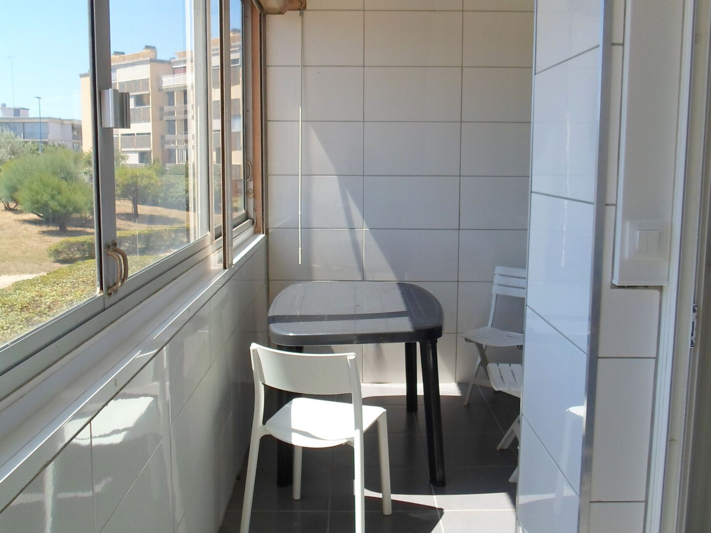 A small balcony with a table and two chairs, a window with a view of buildings, and a tiled wall.