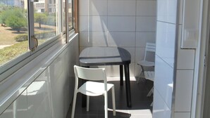 En liten balkong med ett bord och två stolar, ett fönster med utsikt över byggnader och en vägg täckt med kakel.