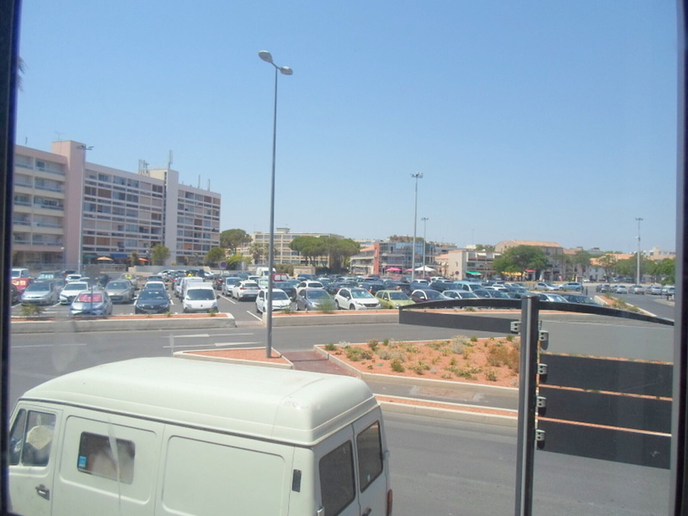 A parking lot with multiple cars and a white van. There are buildings in the background and a clear sky.