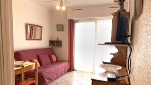 Un espacio habitable pequeño con sofá, una mesita, televisor sobre un mueble de madera y ventilador de techo.