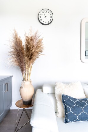 Un sofá blanco con un almohadón blanco mullido y un almohadón azul estampado, un jarrón con plumas de pampa secas y un reloj de pared.