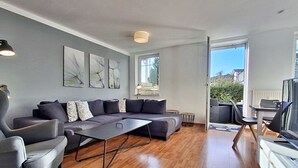 Een moderne woonkamer met een grijze hoekbank, een klein bijzettafeltje en een televisie op een meubel. Er ligt een houten vloer en een groot raam zorgt voor natuurlijk licht.