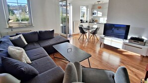 Een moderne woonkamer met een hoekbank, een eettafel met stoelen, een televisie en een keukenhoek.