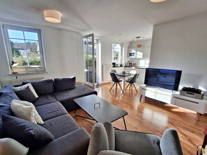 Ein modernes Wohnzimmer mit einem Ecksofa, einem Esstisch mit Stühlen, einem Fernseher und einer Küchenzeile.