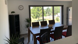 Ein Essbereich mit Holztisch und Korbstühlen, einer Uhr an der Wand und Blick ins Grüne.