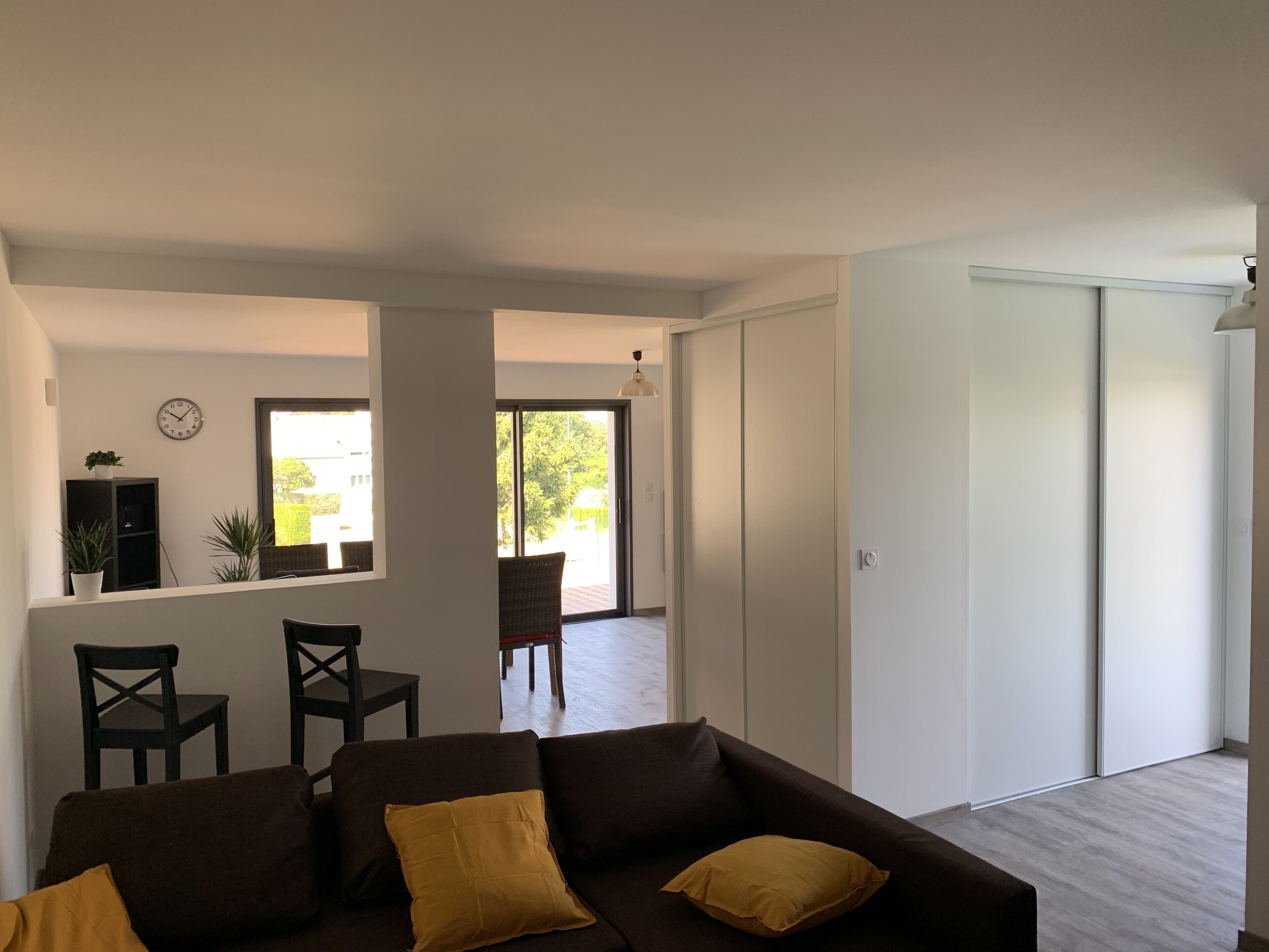 A modern living room with a brown sofa, two yellow cushions, a dining area with chairs, a small plant, and a sliding glass door leading to an outdoor area.