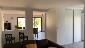 Ein modernes Wohnzimmer mit einem braunen Sofa, zwei gelben Kissen, einem Essbereich mit Stühlen, einer kleinen Pflanze und einer Glastür zum Außenbereich.