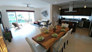 Un vaste espace de vie comprenant un coin repas avec une table pour quatre personnes, une cuisine dotée de meubles sombres et une grande fenêtre offrant une vue sur l’extérieur.