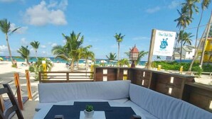 Un espace aménagé près de la plage, avec une table ronde et des chaises, un banc rembourré, et une vue sur la plage et l’océan.