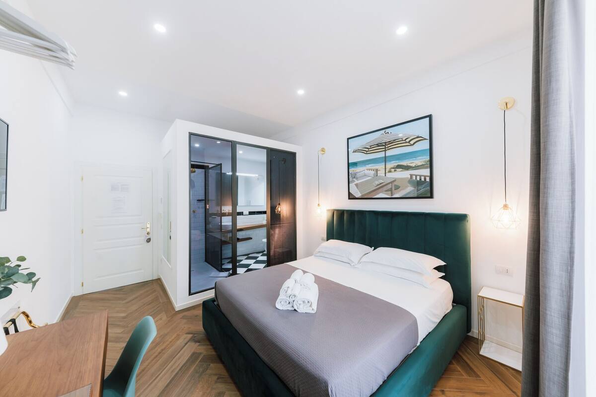 A modern hotel room with a large bed, a desk, and a view of the beach through the window.