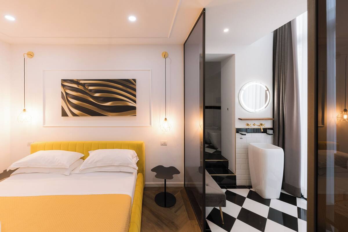A modern hotel room with a yellow headboard bed, a large abstract painting, a round mirror, and a checkered floor.