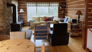 Acogedor interior de cabaña con pared de piedra, techo de madera, un sofá, una silla, una mesa y una estantería.