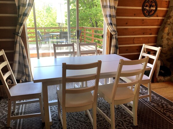 A wooden cabin interior with a dining table and four chairs, a view of the outdoors through large windows, and a stone wall with a decorative metal piece.