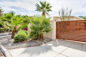 A gated entrance with a palm tree and a small bush, a concrete pathway, and a building with a sloped roof.