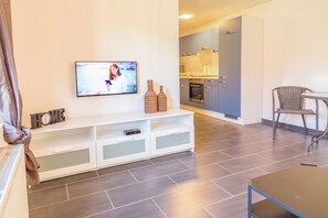 Una sala moderna con un televisor de pantalla plana, un mueble blanco para entretenimiento, una mesa de comedor y una zona de cocina con armarios y una estufa.