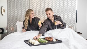 A hotel room with a bed, a tray of food, and two people sitting on the bed. - W Aspen (Aspen)