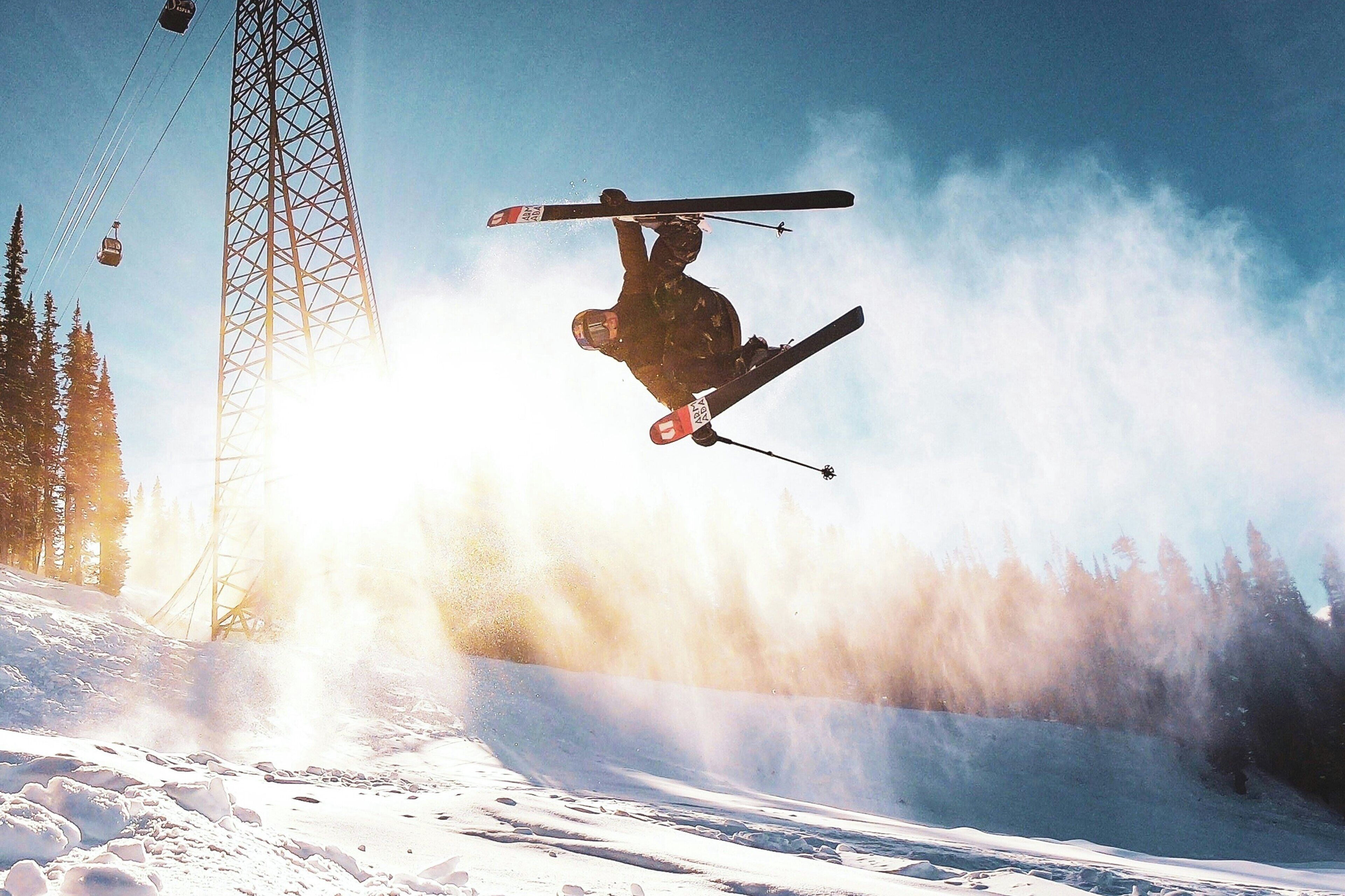A ski jumper in mid-air with skis crossed above their head, a ski lift tower, and a snowy landscape.