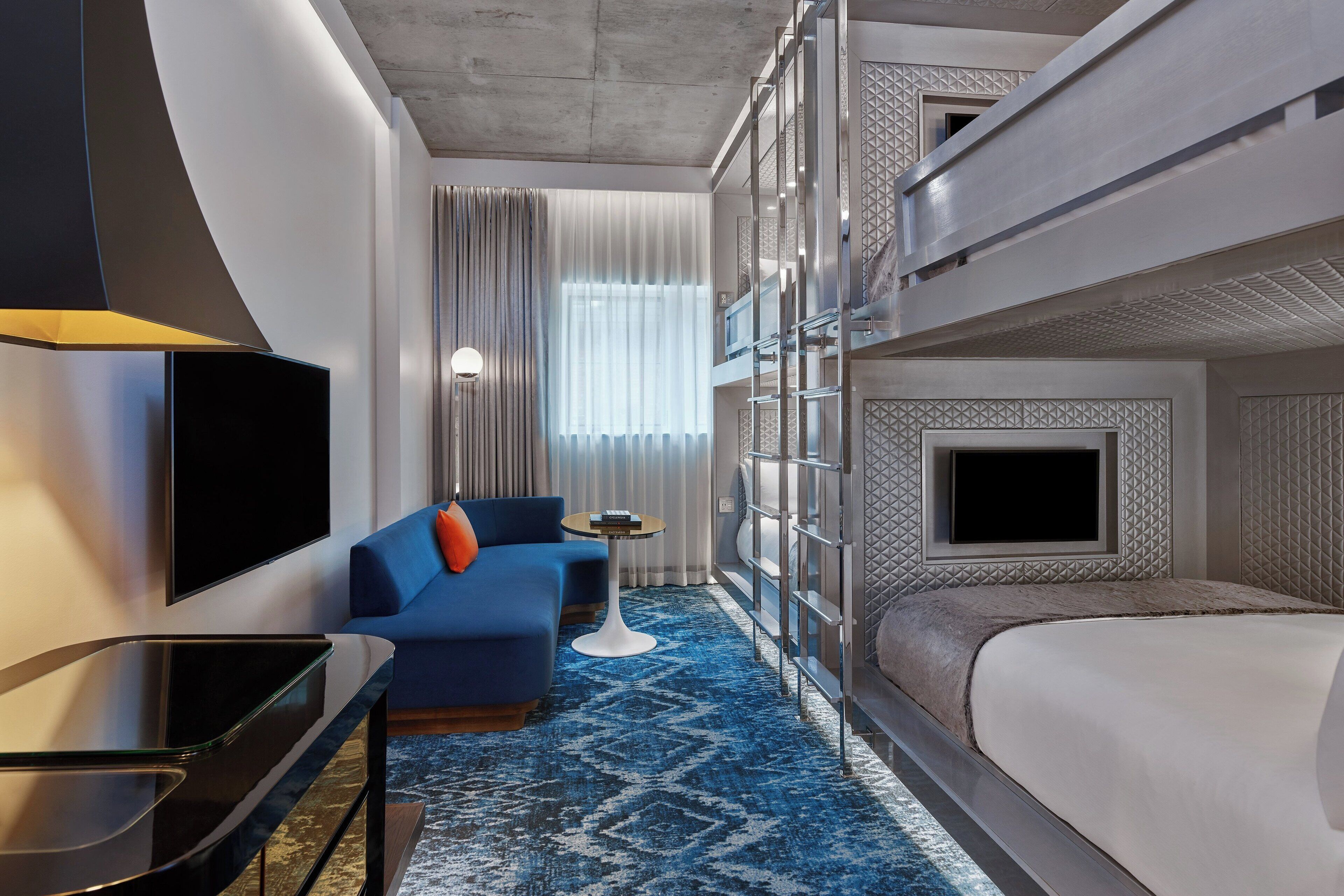 A hotel room with a bunk bed, a flat-screen TV, a sofa, a small table, and a patterned carpet.
