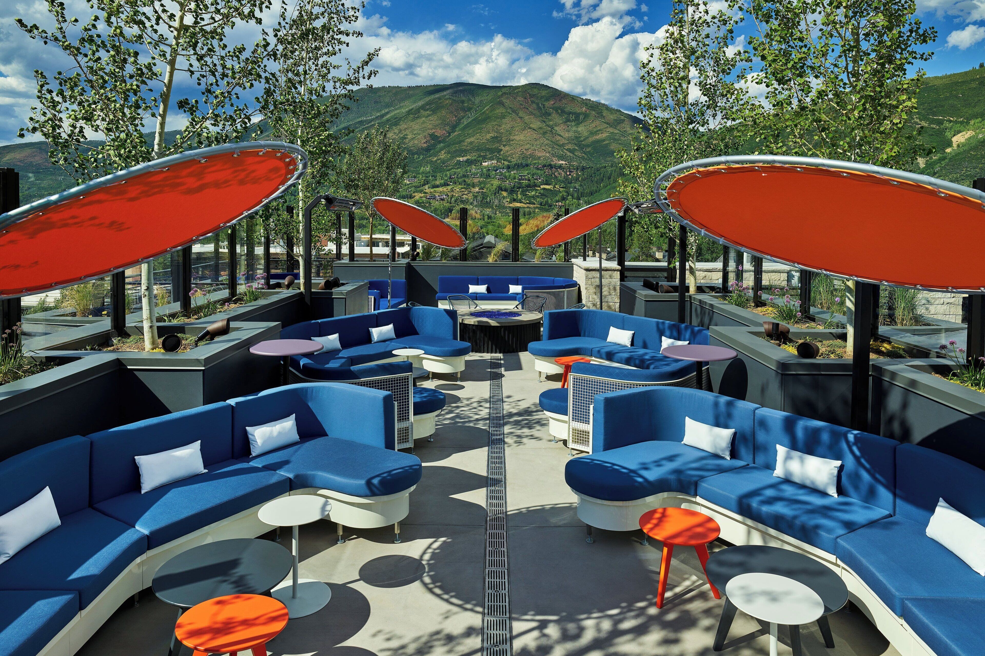 A modern outdoor seating area with blue sofas and red umbrellas, surrounded by mountains.