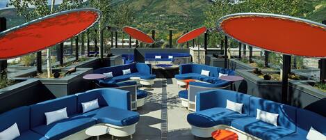 A modern outdoor seating area with blue sofas and red umbrellas, surrounded by mountains.