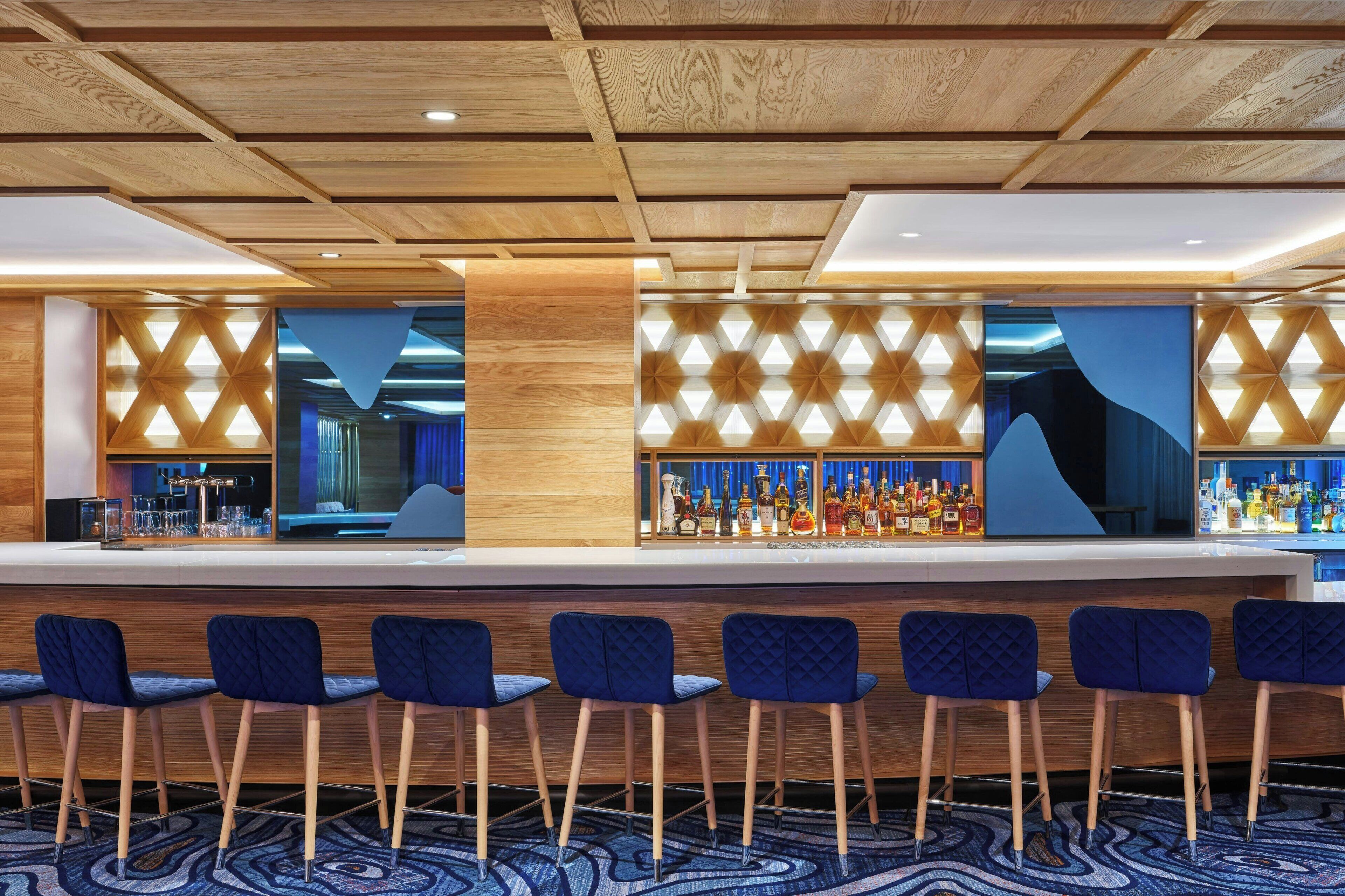 A bar area with a wooden ceiling, blue upholstered bar stools, and a patterned wall design.