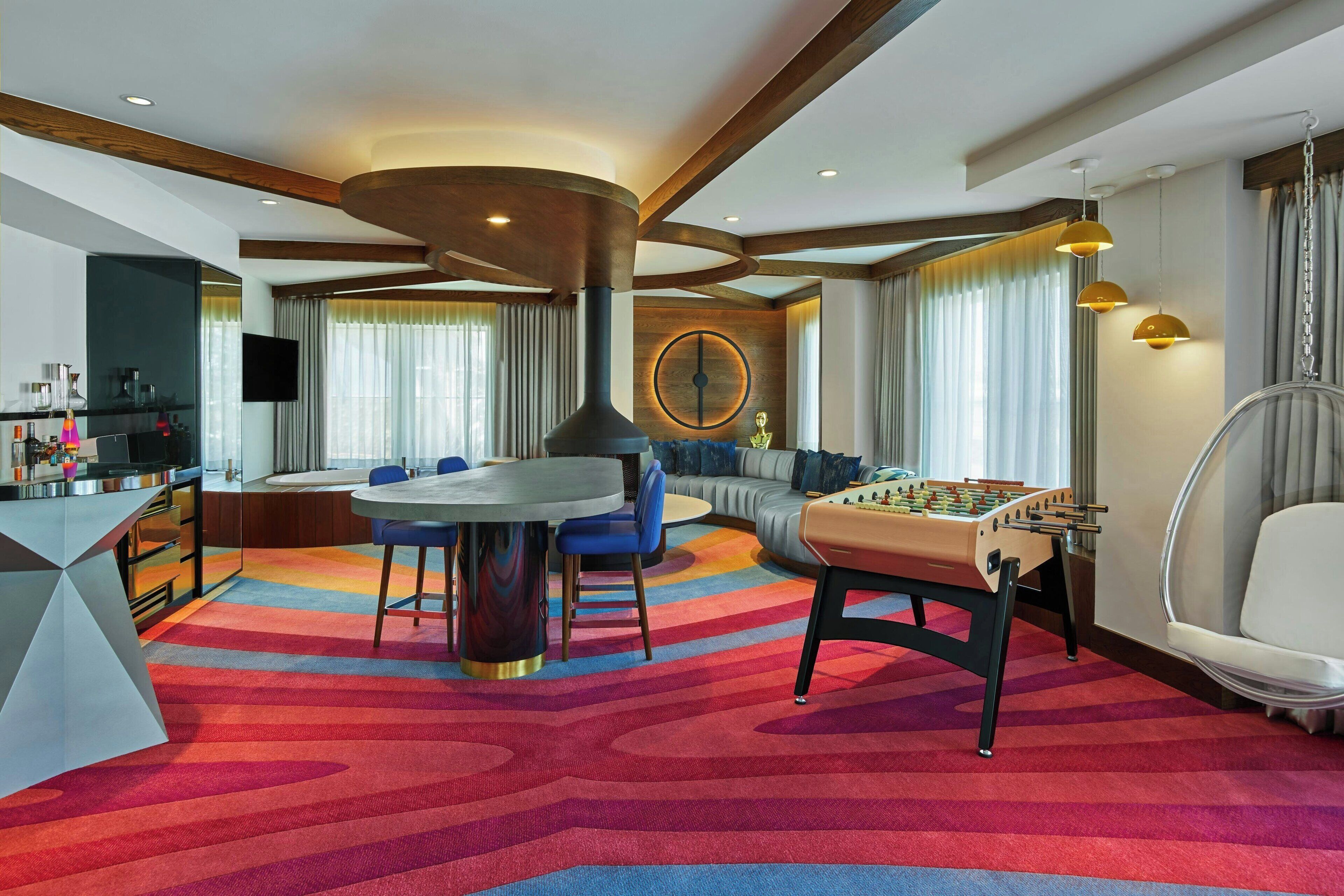 Una habitación de hotel moderna con una mesa redonda de comedor, una mesa de futbolín, un televisor y un ventanal con cortinas.