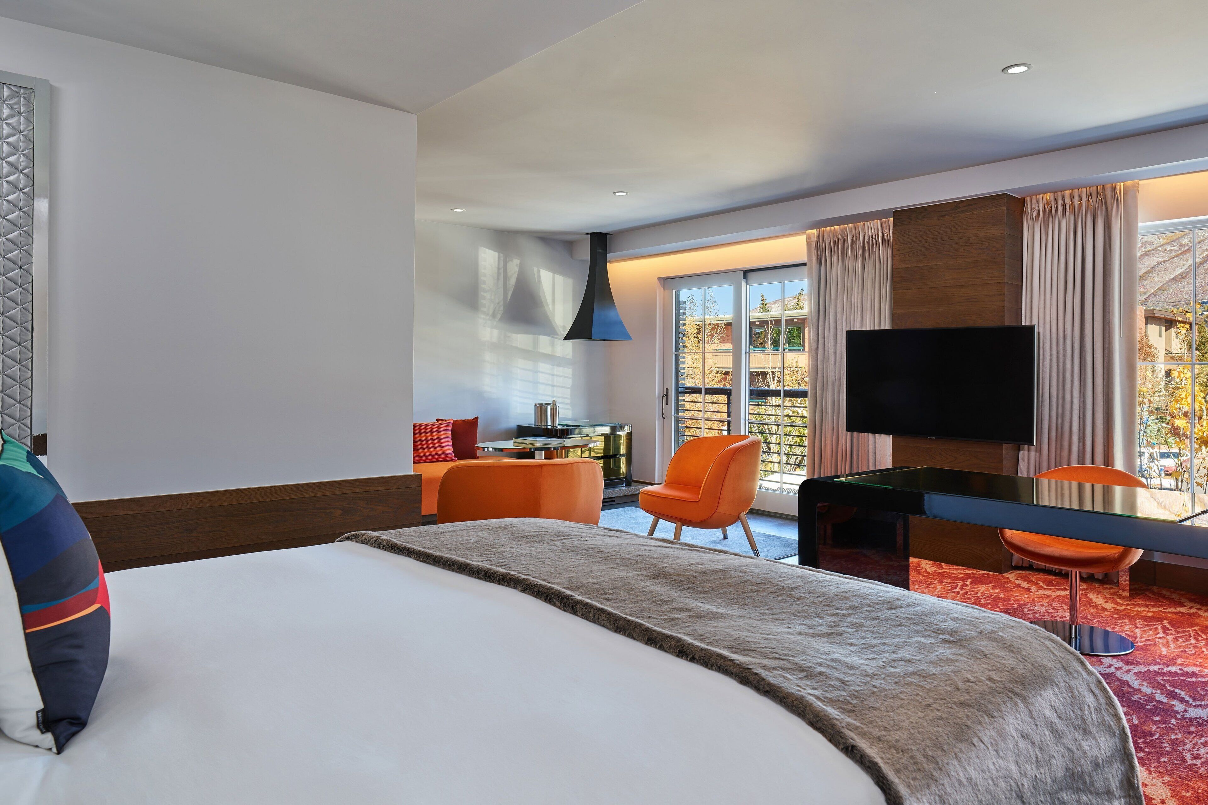 A modern hotel room with a large bed, a flat-screen TV, a dining table, and orange chairs.