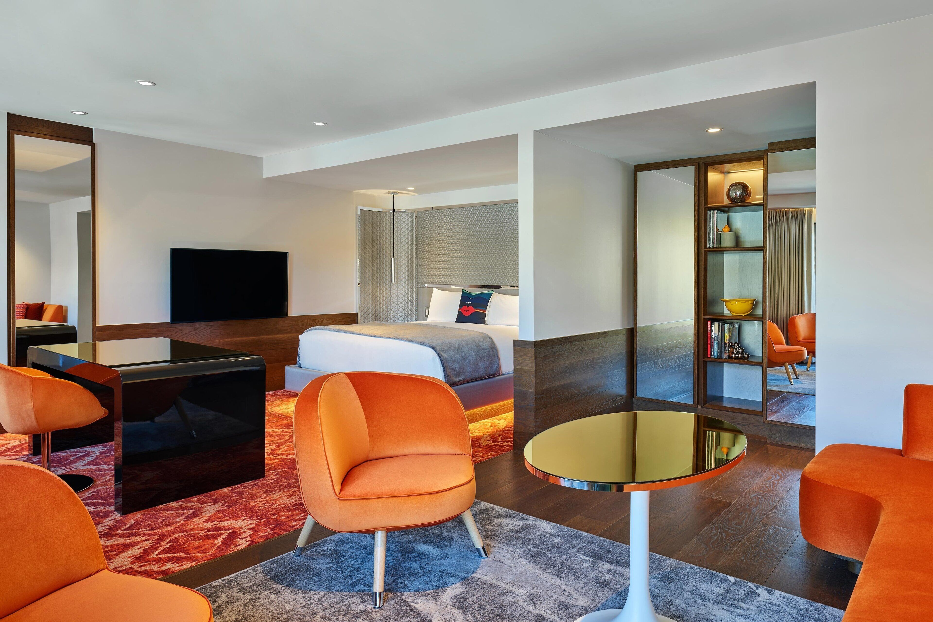 A modern hotel room with a bed, orange armchairs, a glass coffee table, a flat-screen TV, and a bookshelf.