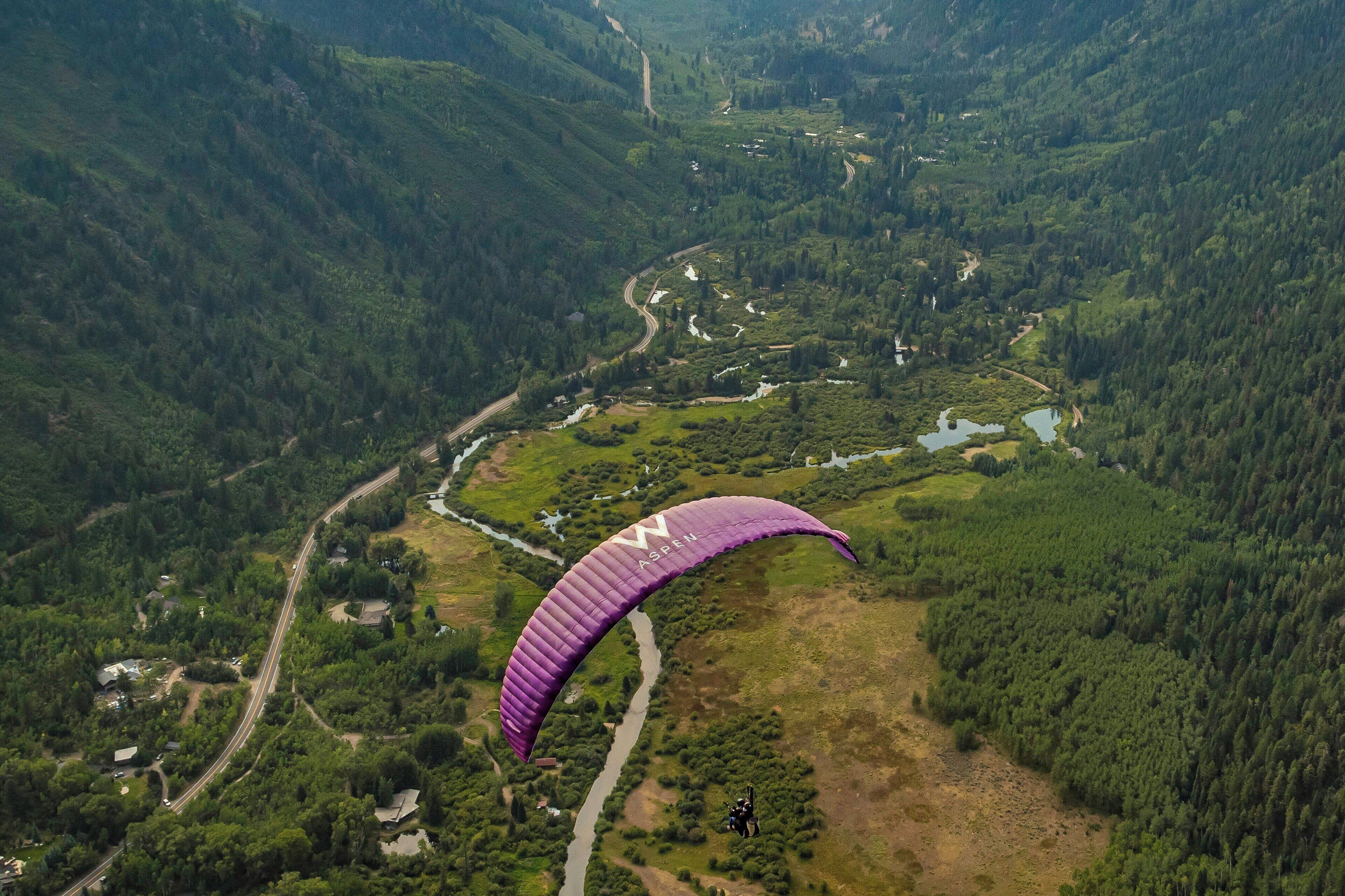 A person paragliding over a mountainous landscape with dense forests and a winding road below.