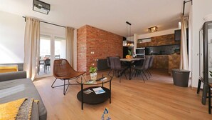 Een moderne woonkamer met een bank, een ronde salontafel en een eethoek met stoelen en een tafel. Er is een keuken met een fornuis en kasten, en een balkon met uitzicht.