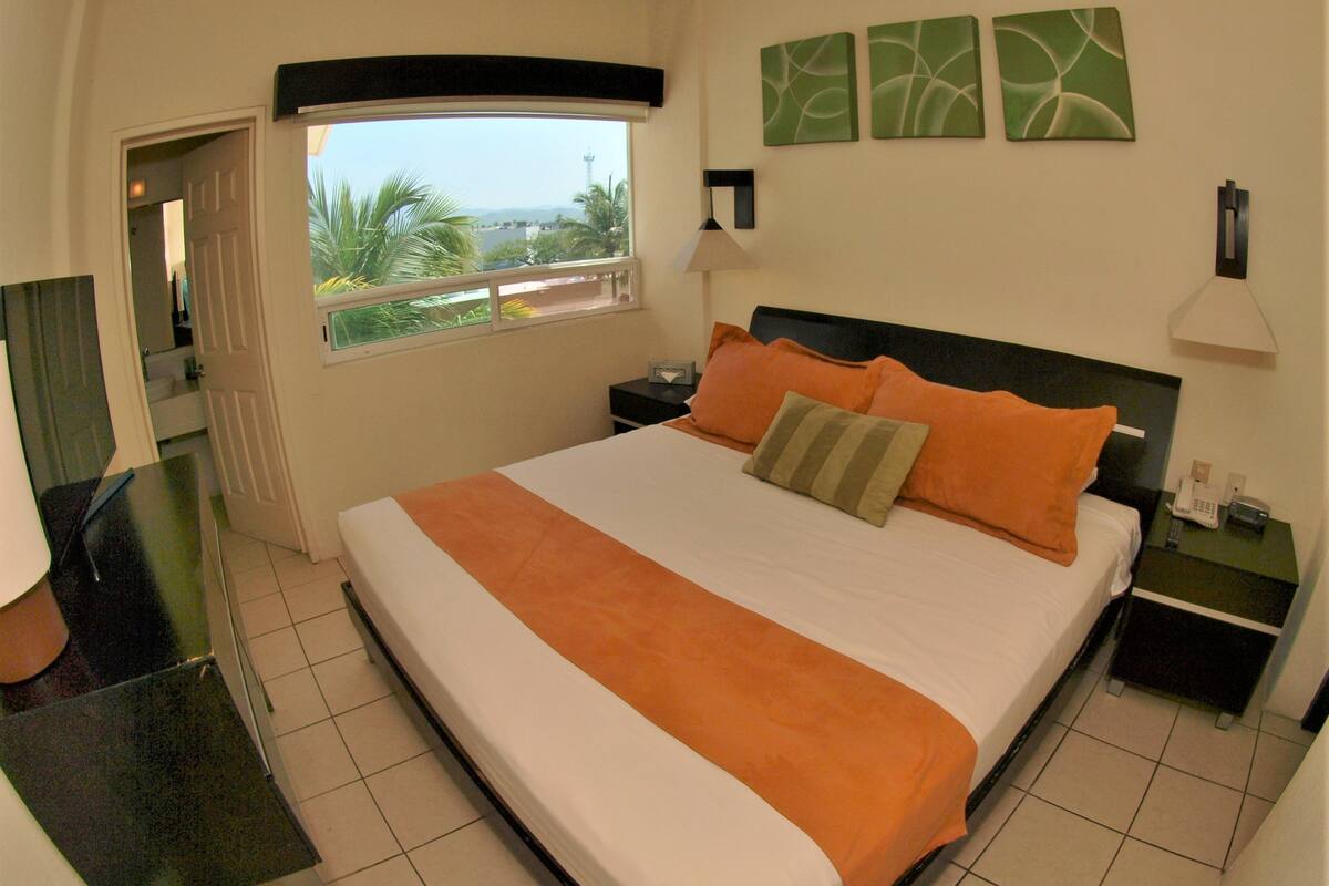 A hotel room with a large bed, a nightstand with a phone, a window with a view of palm trees, and a bathroom door.