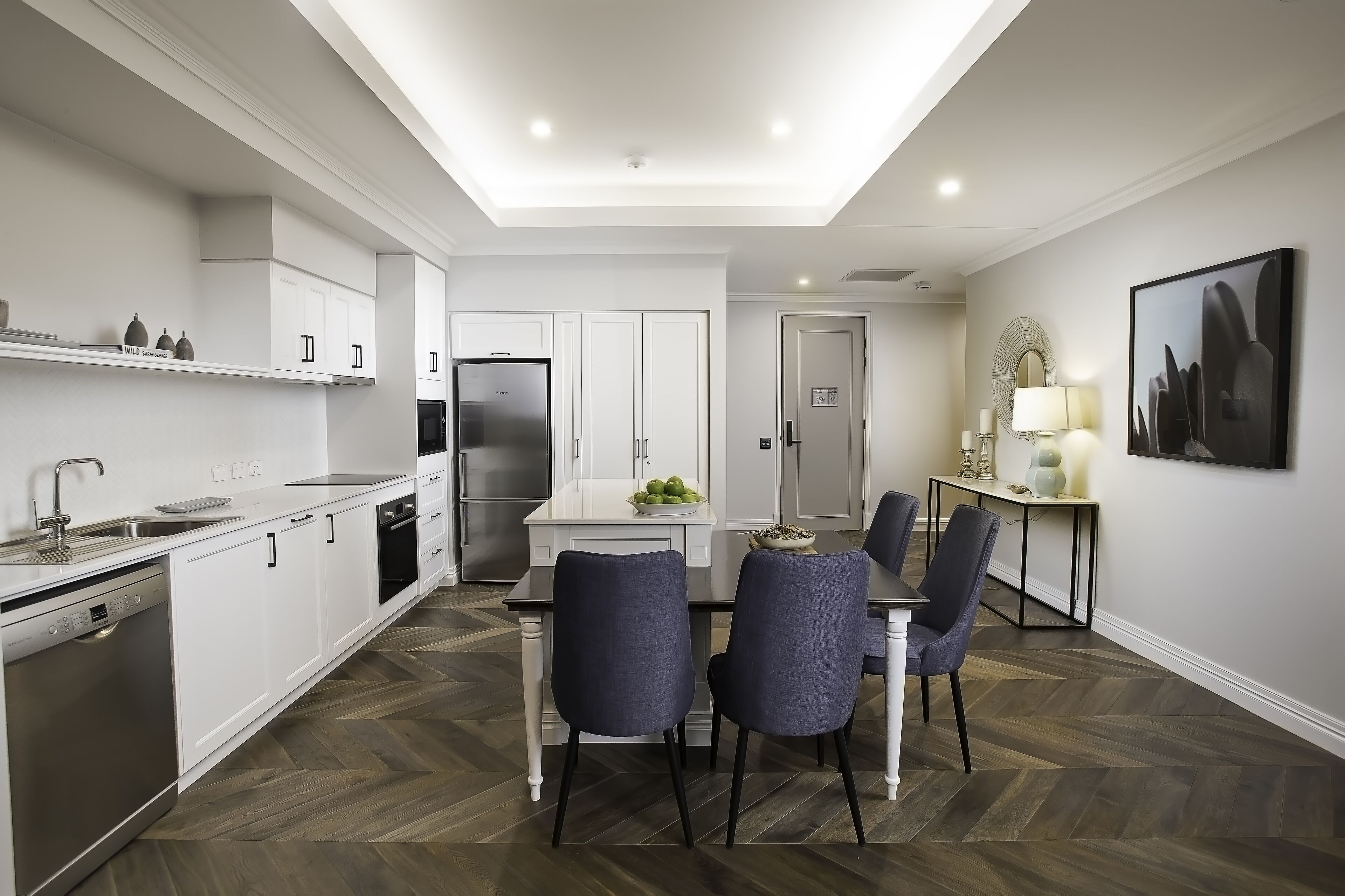 A modern kitchen with white cabinetry, a built-in dishwasher, and a dining area with a table and chairs.