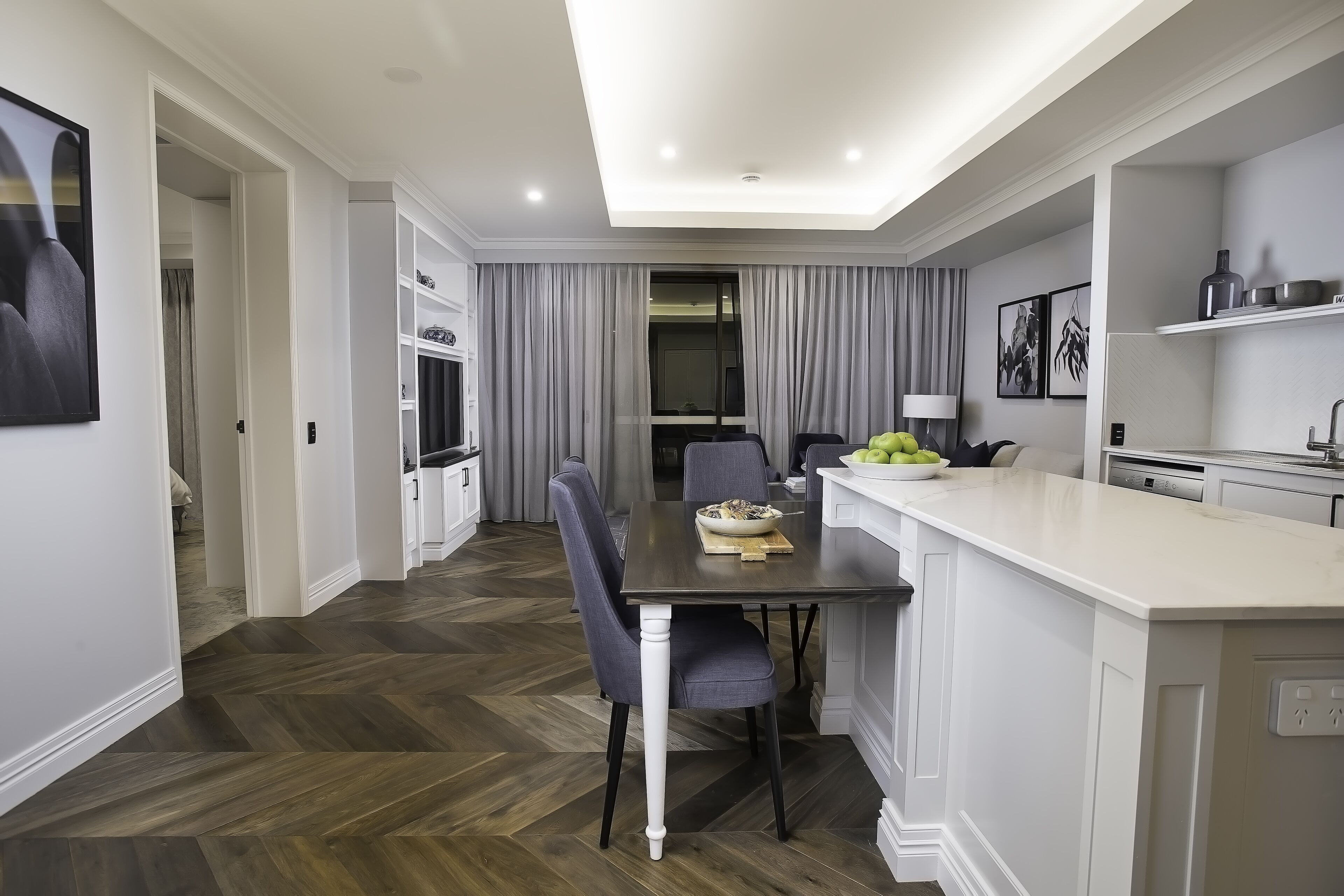 Une cuisine moderne avec un coin repas, dotée d’un parquet en bois disposé en chevrons, de meubles blancs et d’un îlot central sur lequel se trouve un saladier rempli de fruits.
