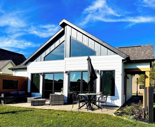 A modern two-story house with a large glass front, outdoor seating area, and a patio.