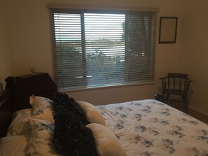 Ein Schlafzimmer mit Bett, Fenster mit Jalousien, einem Sessel und einem kleinen Tisch.