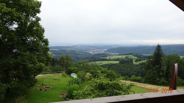 Vista desde un balcón sobre un paisaje verde y exuberante, con un pueblo a lo lejos, un área con césped y una pequeña construcción, además de una fecha que indica 1/07/2019.