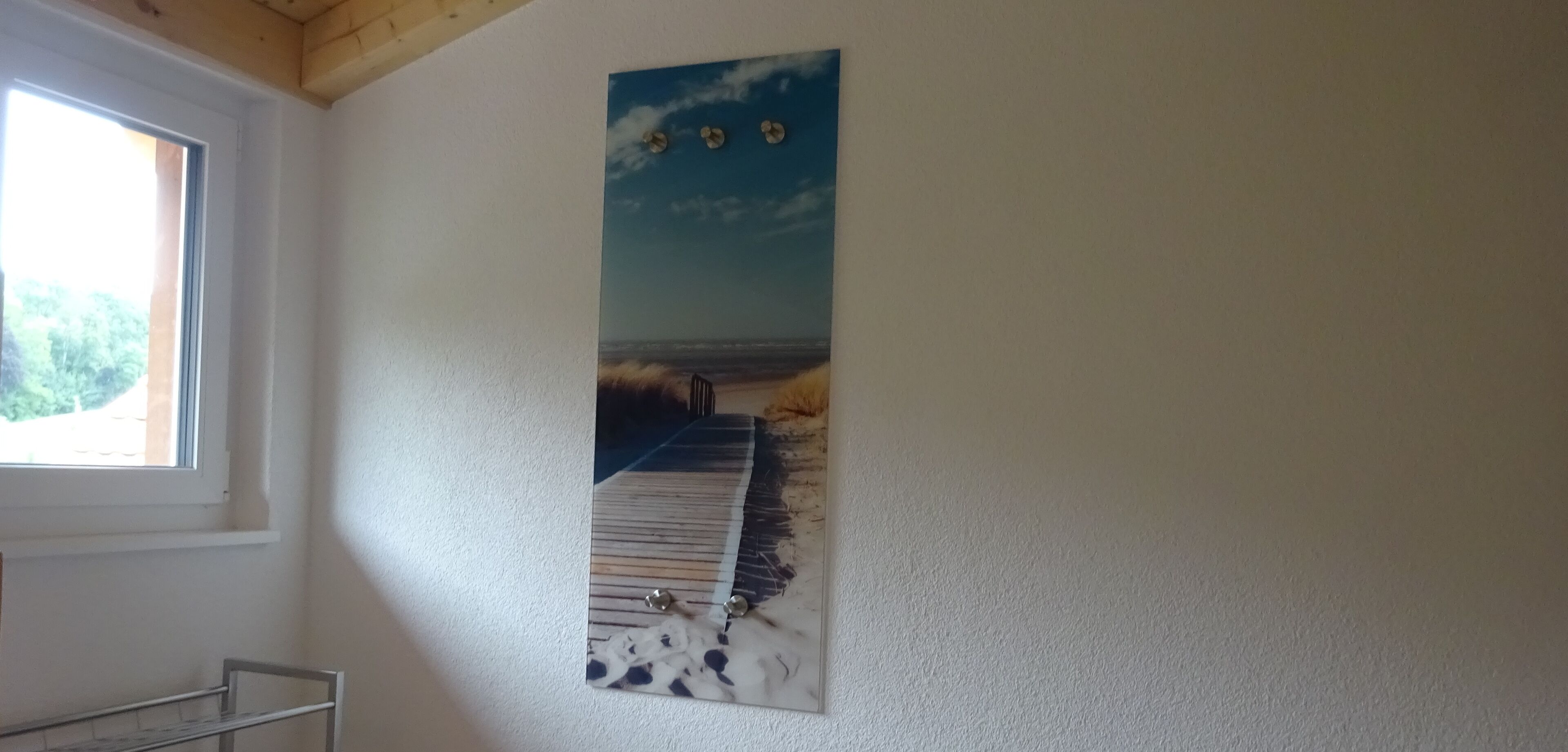 A room with a large painting of a beach scene on the wall, a window with a view of greenery, and a metal shelf.