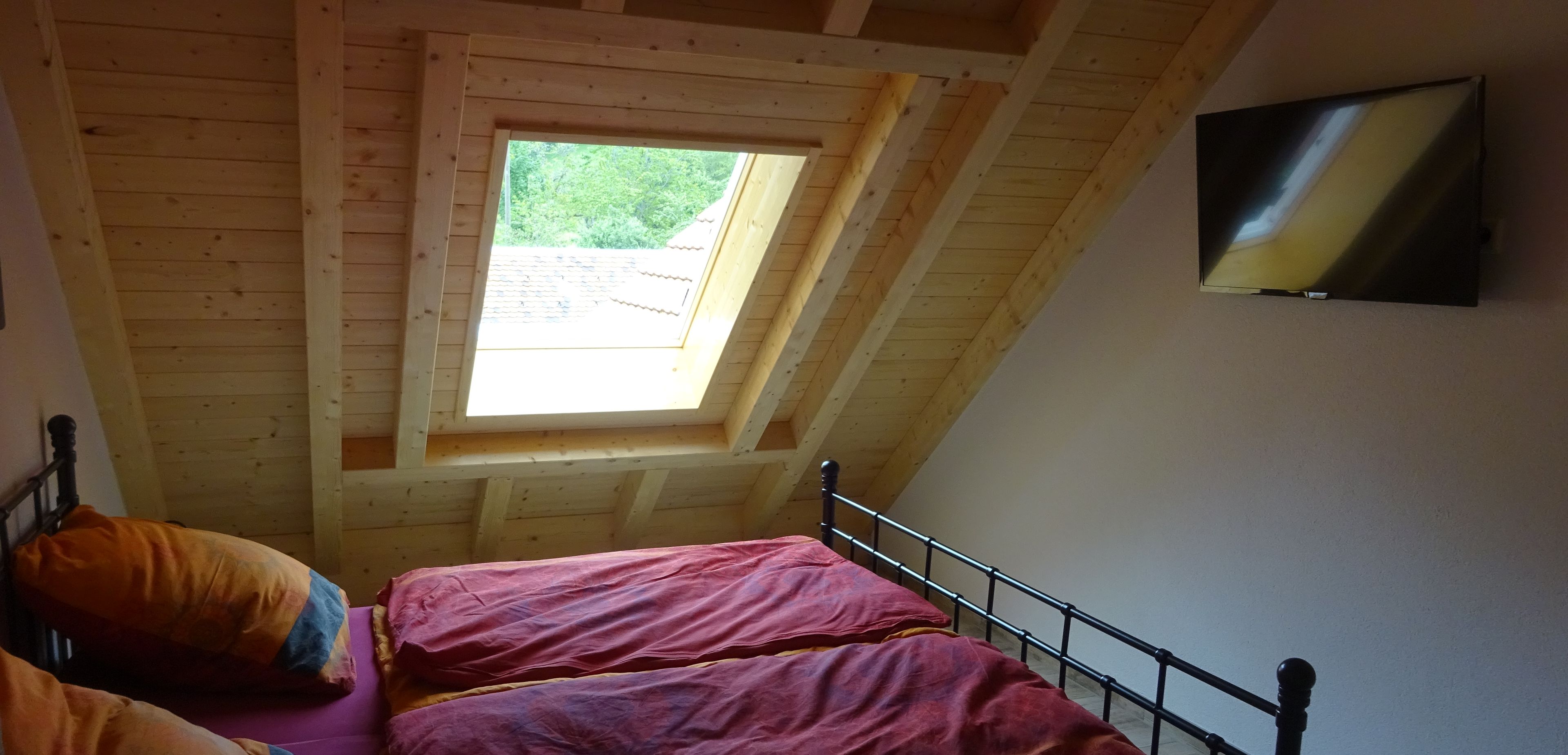 Una habitación con techo inclinado, vigas de madera, dos camas con ropa de cama roja, un televisor en la pared y una ventana con vistas a la vegetación.
