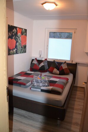 A bedroom with a bed, a window with blinds, a painting on the wall, and a bedside lamp. - Holiday house at Birkenwäldchen - Motzener See 150m (Motzen)