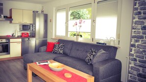A modern living room with a grey sofa, a wooden coffee table, and a kitchen area with a microwave, oven, and refrigerator. - Holiday house at Birkenwäldchen - Motzener See 150m (Motzen)