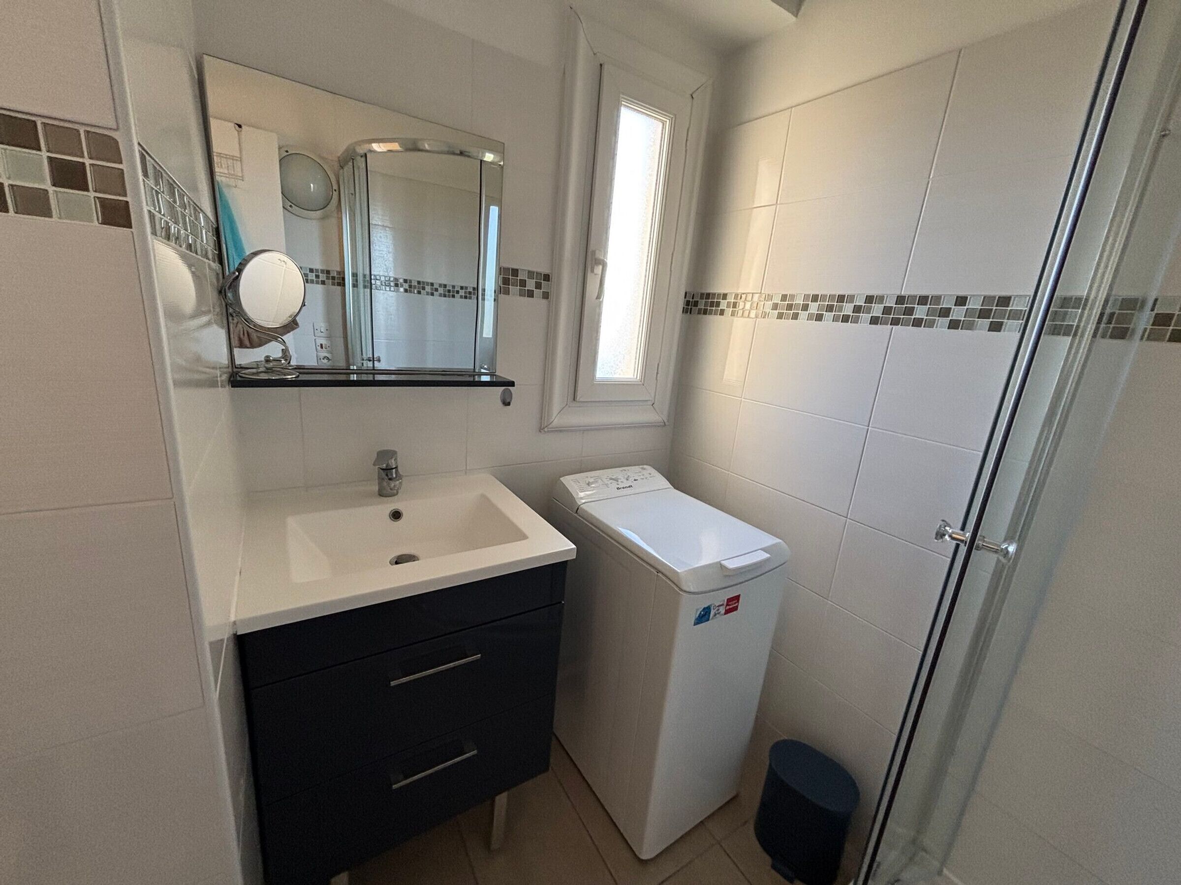 Un baño compacto con lavamanos blanco, un espejo, un ventanal pequeño y una lavadora blanca.