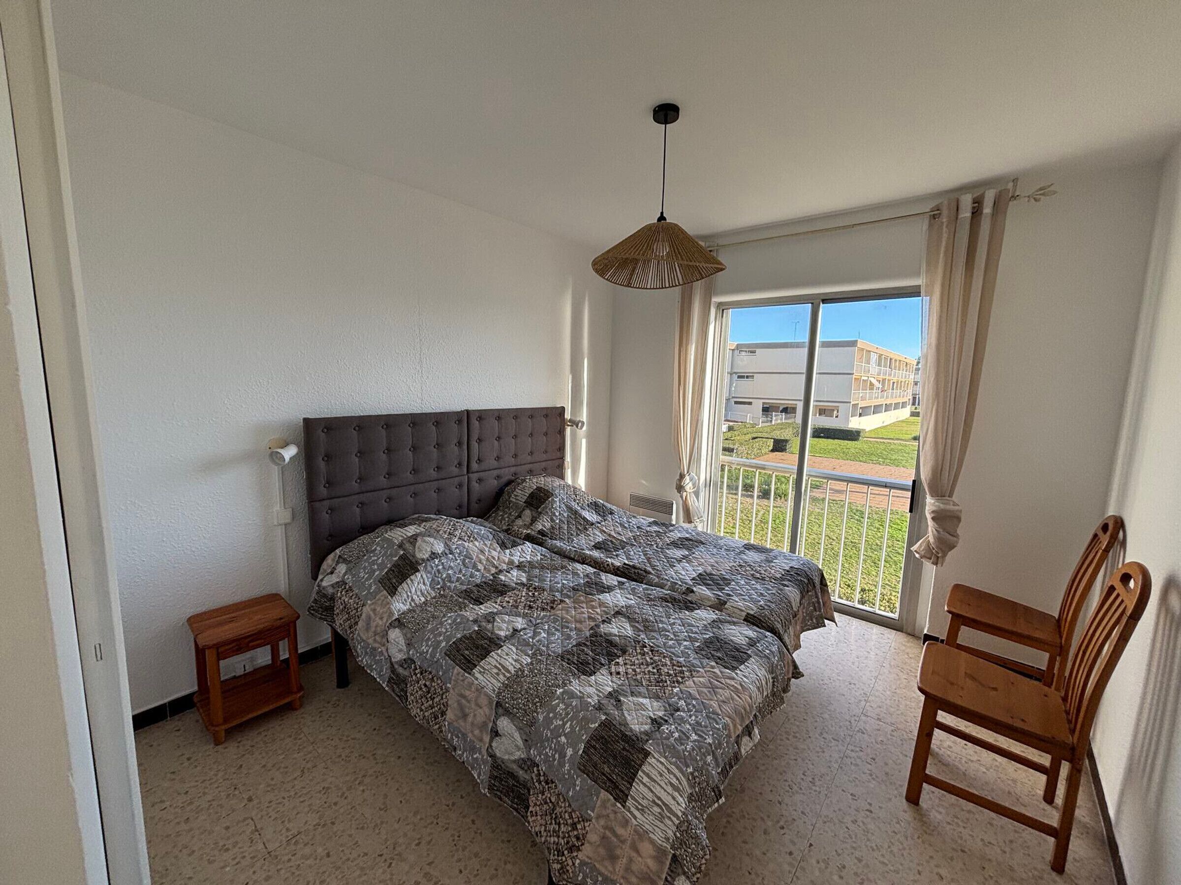 Une chambre à coucher avec un grand lit, une petite table de chevet, une chaise et une vue sur un immeuble par la fenêtre.